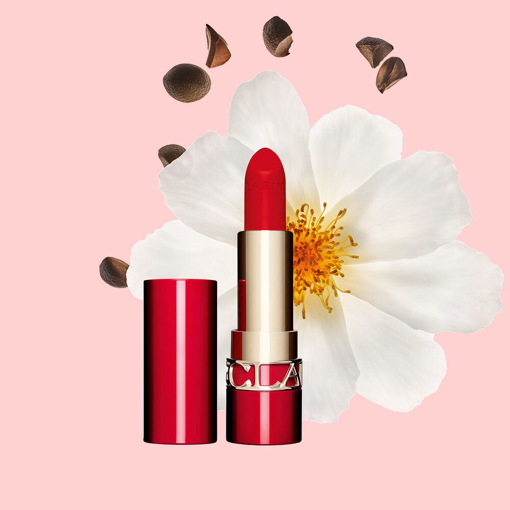 Packshot of Clarins matte Lipstick Joli Rouge in Shade 792 V, placed beside a camellia flower and shea seeds on a pink background.