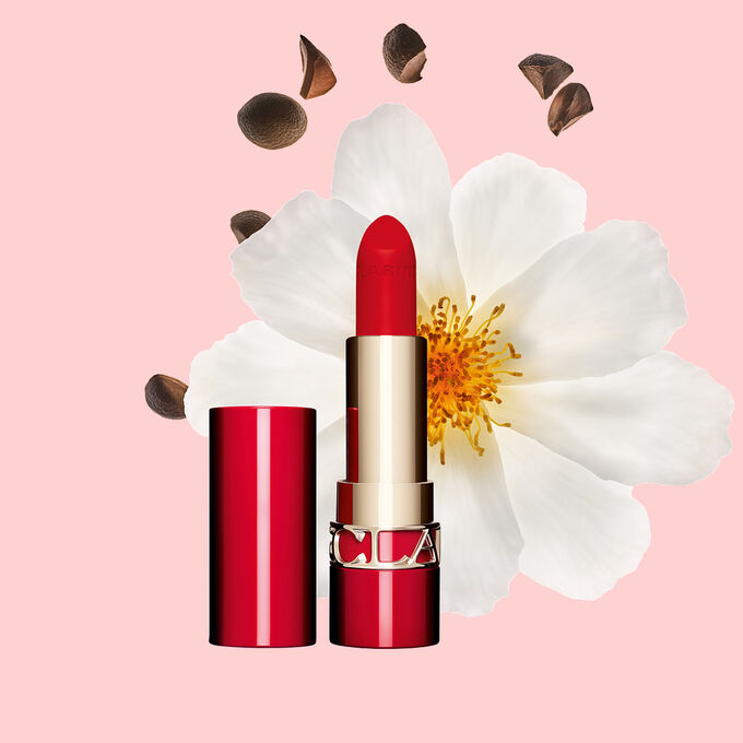 Packshot of Clarins matte Lipstick Joli Rouge in Shade 792 V, placed beside a camellia flower and shea seeds on a pink background.