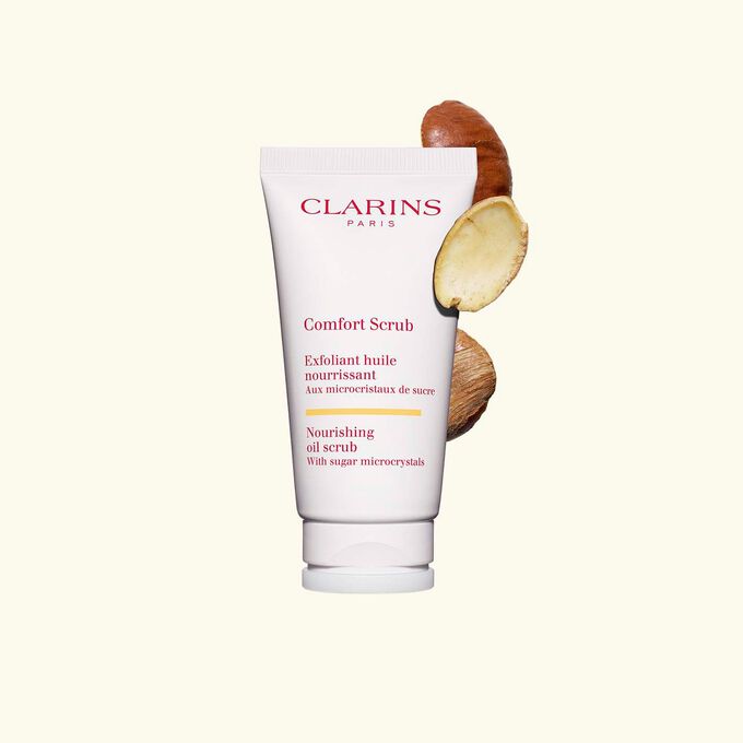 Packshot of Clarins face and lips exfoliant Comfort Scrub tube next to some wild mango seeds on a beige background