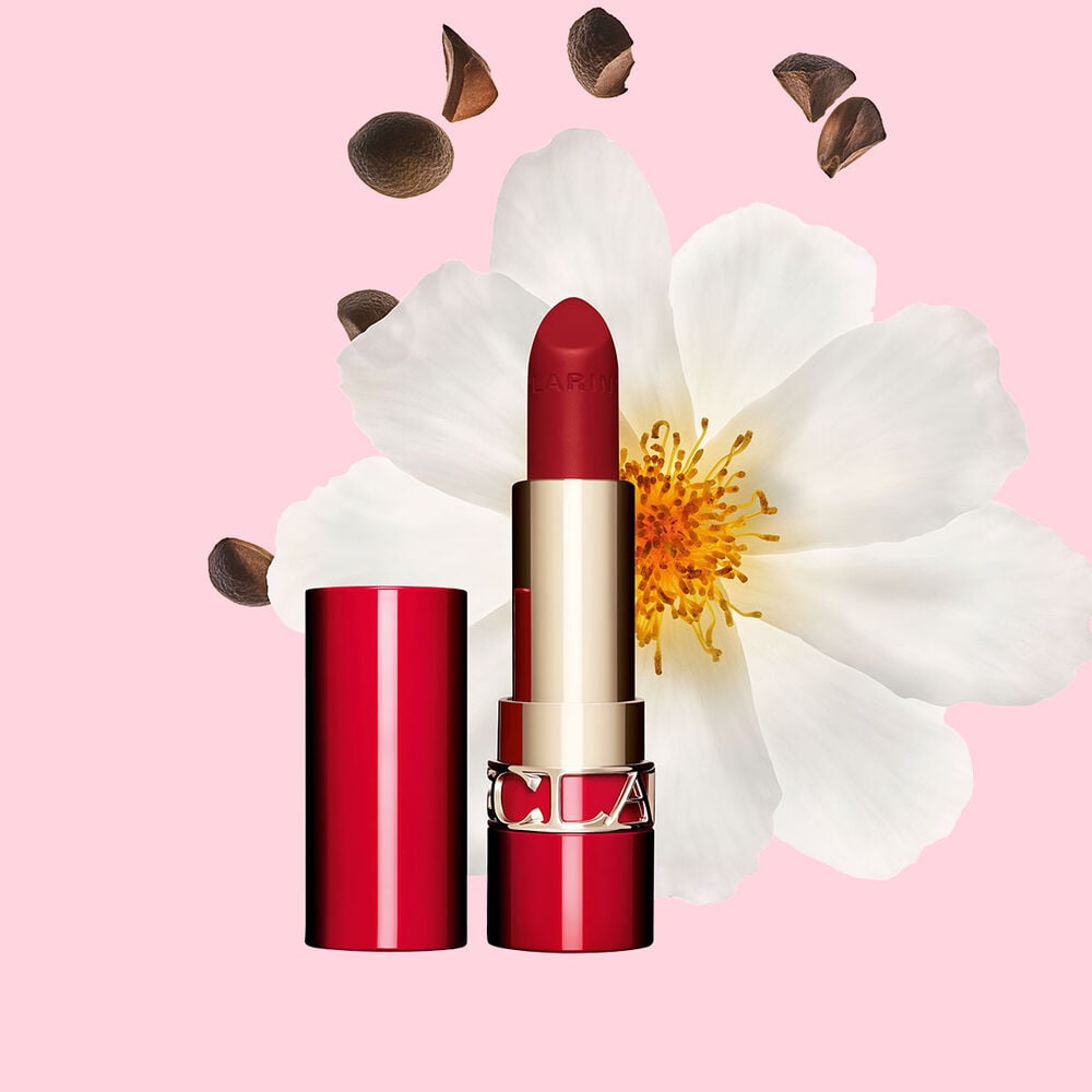 Packshot of Clarins matte Lipstick Joli Rouge in Shade 793 V, placed beside a camellia flower and shea seeds on a pink background.